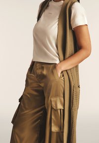 Chaleco largo de punto beige sobre una camiseta blanca con el logo dorado "FREDDY", acompañado de pantalones anchos de satén color caqui con bolsillos laterales.