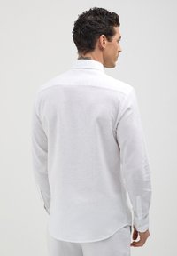 Mann mit kurzen lockigen Haaren, der ein langärmliges weißes Leinenhemd trägt, von hinten vor einem einfarbigen hellen Hintergrund gesehen.