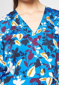 Bluse mit blauem Blumenmuster, V-Ausschnitt, kurzen Ärmeln und einer Mischung aus weißen, gelben und lila Blumen sowie grünen Blättern auf glattem Stoff.