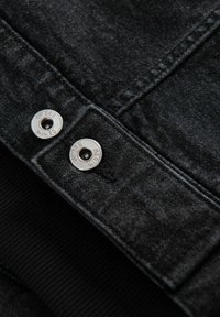 Giacca in denim nera con bottoni a pressione argentati e polsini neri a coste. La superficie presenta una finitura morbida e strutturata con sottili variazioni.