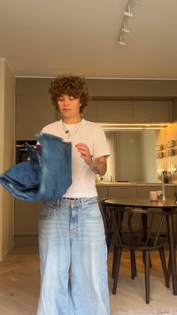 Jeans din denim de culoare albastră deschisă, cu croială largă, fiind ținuți de cineva, având o textură ușor distresată și un design clasic cu cinci buzunare. Se poartă un tricou simplu alb.