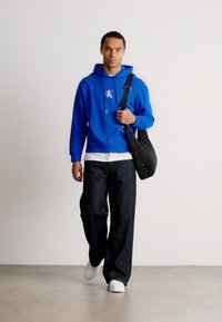 Benn járó férfi kék kapucnis pulóverben, fehér sportcipőben, fekete bő nadrágban, és egy fekete válltáskával.