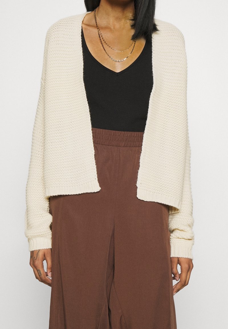 Knit cream cardigan with an open front, textured fabric, wide sleeves, worn over a black fitted top and brown elastic-waist pants.