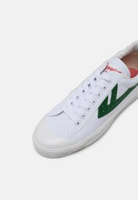 White canvas sneaker featuring a green leather accent and red branding. Rounded toe, flat laces, and textured white rubber sole.