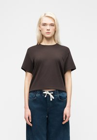 Νεαρή γυναίκα με μακριά ξανθά μαλλιά, φορώντας ένα φαρδύ σκούρο καφέ μπλουζάκι και σκούρα μπλε παντελόνια με κορδόνι, μπροστά σε απλό φόντο.