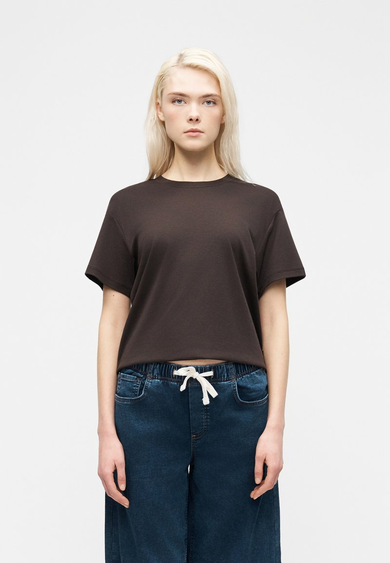 Νεαρή γυναίκα με μακριά ξανθά μαλλιά, φορώντας ένα φαρδύ σκούρο καφέ μπλουζάκι και σκούρα μπλε παντελόνια με κορδόνι, μπροστά σε απλό φόντο.