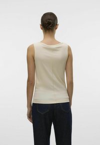 Beżowy top na ramiączkach o okrągłym dekolcie i pionowym szwem z tyłu, zestawiony z ciemnoniebieskimi spodniami dżinsowymi. Gładka tekstura.