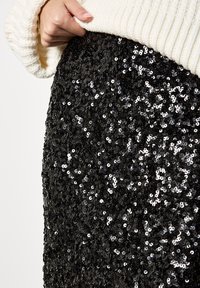 Schwarzer Paillettenrock mit einer dichten Anordnung runder Pailletten, die eine schimmernde Textur zeigen. Ein cremiger, gerippter Pullover ist teilweise sichtbar.