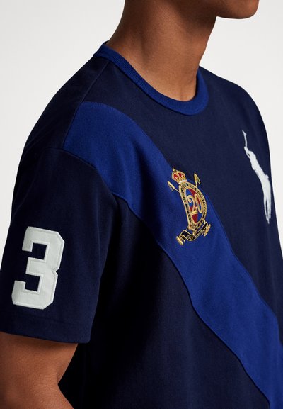 Tmavě modré polo tričko s kontrastním modrým diagonálním panelem, vyšitým erbem a velkým bílým číslem "3" na rukávu.