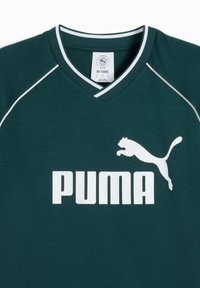 Tamsiai žalia sportinė marškinė su V formos iškirpimu, baltais akcentais ir dideliu baltu "PUMA" logotipu su šokančio pumos piešiniu. Tekstūruotas audinys.