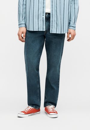 Inimene, kes kannab tumelsiniseid sirge lõikega teksaseid, punaseid lõuendist tossusid valgete paeltega ja helesinist triibulist nööpidega särki.