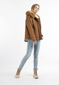 Brun isoleret jakke med pelsforet hætte, lynlås lukning og to sidelommer. Båret over lyseblå slidte jeans og beige høje sneakers.