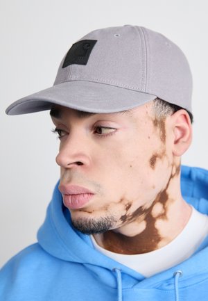 Gray baseball cap with a black textured logo patch, featuring a curved brim and eyelet details. Worn with a light blue hoodie.
