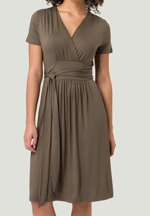 Femme portant une robe cache-cœur olive mi-longue à manches courtes avec une ceinture nouée et une jupe plissée, debout devant un fond uni.