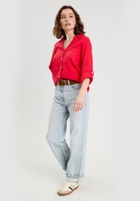 Rotes Leinen-Hemd mit Knopfleiste und hochgekrempelten Ärmeln, kombiniert mit hellblauen, locker geschnittenen Jeans und beigen Sneakers mit braunen Akzenten.