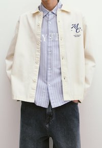 Cream-colored cotton shirt jacket with a button-front, worn over a blue striped long-sleeve shirt. Dark jeans visible below.
