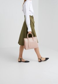 Borsa a tracolla in pelle rosa chiaro con forma rettangolare strutturata, dotata di due manici superiori e dettaglio della fascia frontale; il modello indossa una gonna verde oliva e ballerine nere.