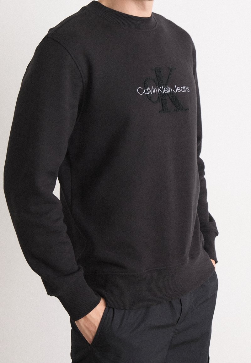 Czarny sweter z okrągłym dekoltem, wykonany z bawełny, z haftowanym napisem "Calvin Klein Jeans" w kolorze białym oraz wyraźnym logo "CK".