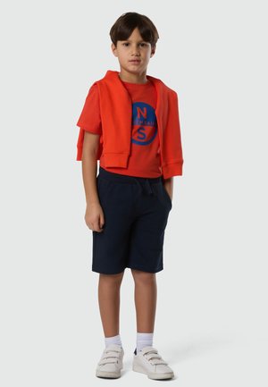 Junge in orangem T-Shirt und Sweatshirt über den Schultern, marineblaue Shorts, weiße Socken und weiße Turnschuhe, der vor einem schlichten Hintergrund steht.