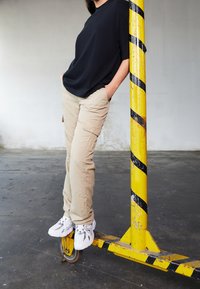Černé oversized tričko, béžové cargo kalhoty s kapsami a bílé tenisky. Stojí vedle sloupu se žluto-černými pruhy.