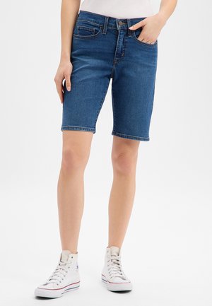Femme portant un short en jean bleu mi-long et des baskets blanches montantes, debout avec une main dans la poche.