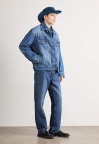 Denimjakke med vertikale blå striper, to brystlommer og knappelukking. Kombinert med blå jeans og en matchende blå denimhatt.
