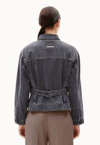 Schwarze Denimjacke mit einem taillierten Gürtel, Seitentaschen und einem rechteckigen Logo-Patch im oberen Rückenbereich. Abgeschlossen mit sichtbaren Nähdetails.