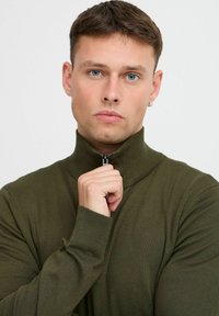 Olibnozeleni pleteni pulover z visokim ovratnikom in pol-zadrgo. Ima rebraste manšete in mehak občutek, oblikovan kroj.