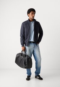 Marine bomberjakke, lys blå t-shirt, lys blå jeans, sort læder duffeltaske, sorte sneakers med hvide striber. Neutral baggrund.