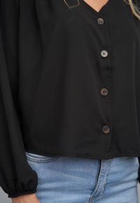 Camisa negra de manga larga con escote en V y botones marrones. Hecha de tela ligera, con un ajuste suelto y un dobladillo corto.