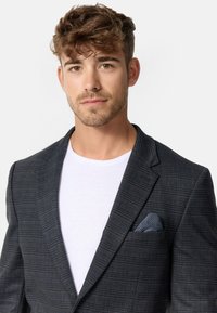 Dunkel gestreifter Blazer mit Revers, strukturiertem Stoff und einem blau-weißen gemusterten Einstecktuch, getragen über einem weißen T-Shirt.