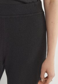 Czarne dzianinowe spodnie o miękkiej teksturze i elastycznym pasie, zbliżenie na materiał oraz dłoń opadającą na boku.