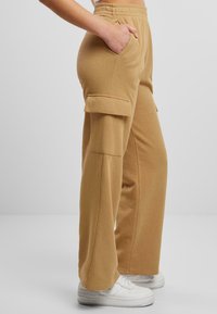 Beige Cargo-Hosen aus weichem Material mit lockerer Passform, Seitentaschen und Klappentaschen mit Knopf an den Oberschenkeln, kombiniert mit weißen Sneakern.