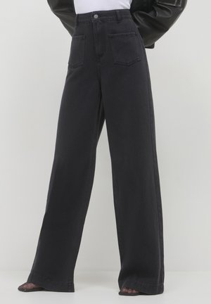 Hochtaillierte, weite, schwarze Jeans mit Fronttaschen, getragen mit schwarzen Mesh-Pumps mit spitzem Zeh und einem weißen, in die Hose gesteckten Oberteil.