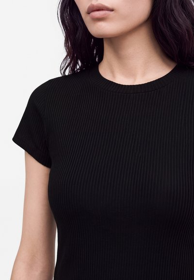 Femme aux cheveux foncés portant un haut noir côtelé à manches courtes, montrant le cou, les épaules et le haut de la poitrine sur un fond uni.