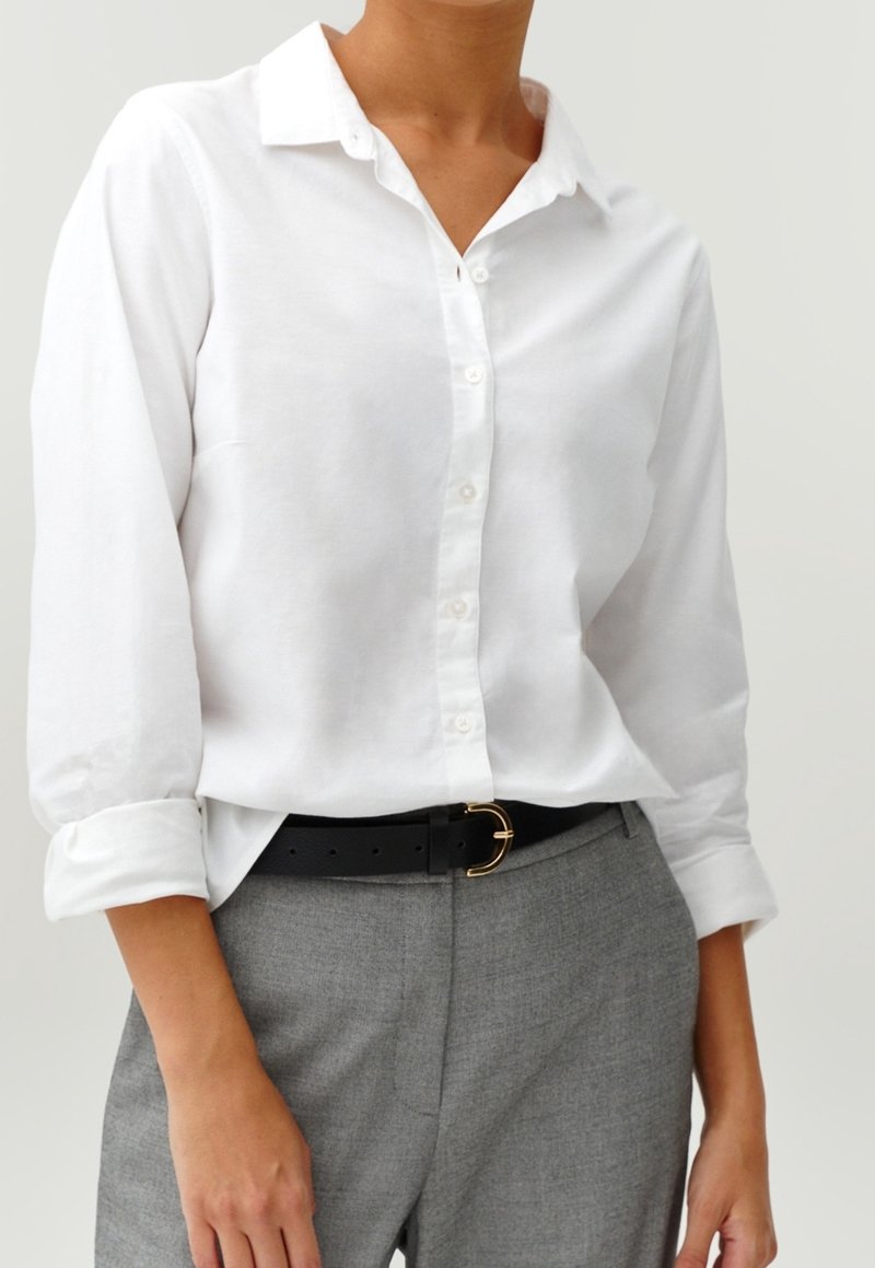 Chemise blanche à manches longues, en tissu texturé, avec un col, portée avec un pantalon gris et une ceinture noire avec une boucle dorée.