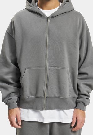 Man wearing a gray zip-up hoodie over a white shirt with matching gray pants, hands relaxed at sides.