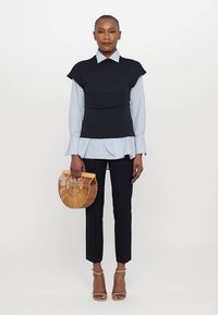 Zwarte aansluitende top over een lichtblauw gestreept shirt, zwarte op maat gemaakte broek, tan hakken en een ronde bamboe handtas. Soepele texturen en gelaagde look.