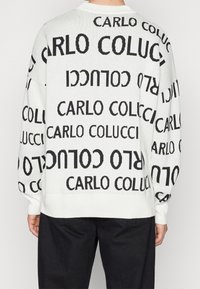 Jersey de punto blanco con un patrón de texto negro repetido "CARLO COLUCCI". El diseño incluye hombros caídos y puños acanalados.