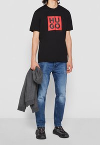 Czarny T-shirt z czerwonym kwadratowym logo z napisem "HUGO", w połączeniu z niebieskimi dżinsami i czarnymi trampkami. Szary sweatshirt trzymany jest w lewej ręce.