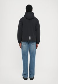 Svart vadderad jacka med huva, med en randig logodetalj på baksidan. Modellen bär ljusblå jeans och svarta skor.