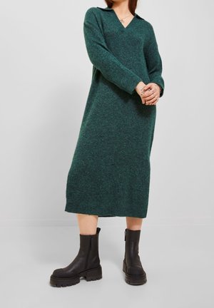 Vrouw draagt een donkergroene, lange mouwen, gebreide midijurk met een kraag en zwarte, stevige enkellaarzen, staand met gevouwen handen.