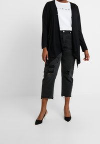 Kobieta w czarnym otwartym kardiganie, białej koszulce z grafiką, podartych czarnych krótkich dżinsach i czarnych szpilkach stoi.