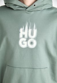 Sudadera verde con una textura suave, que presenta un gráfico blanco "HUGO" con acentos de llamas. Incluye un bolsillo tipo canguro y capucha ajustable.