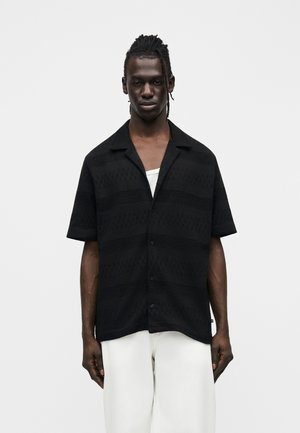 Homme aux cheveux tressés, portant une chemise noire texturée à manches courtes boutonnée par-dessus un haut blanc et un pantalon blanc, debout devant un fond uni.