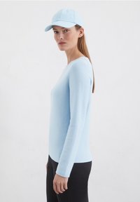 Helles blaues Langarmoberteil mit gerippter Textur, kombiniert mit schwarzen Leggings und einer passenden hellblauen Baseballmütze, von der Seite betrachtet.