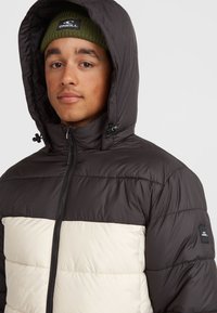 Gewatteerde jas met een zwart bovenstuk en een crèmekleurig onderstuk, voorzien van een capuchon en ritsluiting. Inclusief logopatch op de mouw.