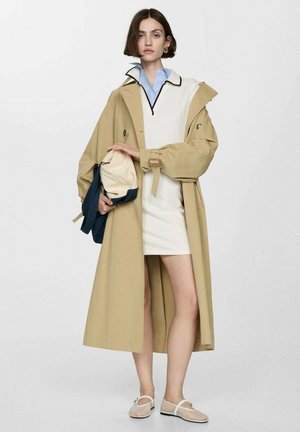 Cappotto beige con maniche ampie, vita stretta e tasche grandi, indossato su un vestito bianco e abbinato a scarpe chiare.