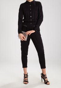 Jumpsuit noir avec un devant à boutons, deux poches poitrine, une taille élastique, associé à des talons strappy noirs et une pochette métallique.