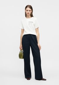 Γυναίκα με λευκό T-shirt με στάμπα "weekend", μαύρο παντελόνι φαρδιάς γραμμής, καφέ παπούτσια, κρατώντας μικρή τσάντα ώμου σε ελαιόχρωμο, στέκεται σε λευκό φόντο.